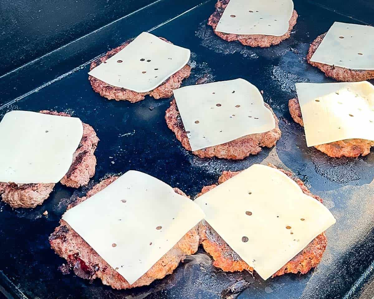 Slices of cheese melting over venison smash burger patties on a flat-top griddle.
