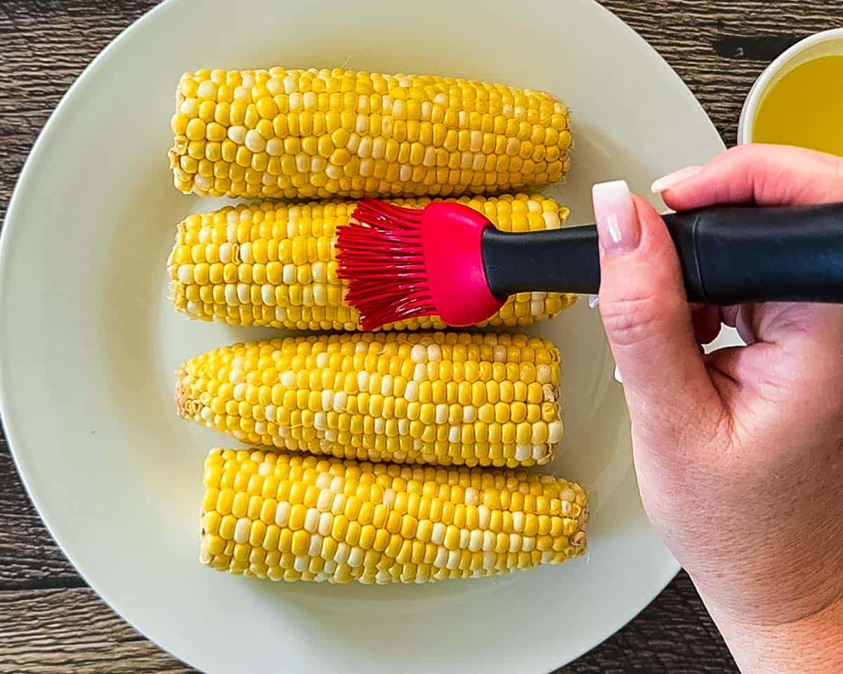 Hand brushing oil onto four ears of corn on a white plate before air frying.