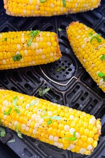 Four ears of air fryer corn on the cob seasoned with black pepper and fresh herbs in an air fryer basket.