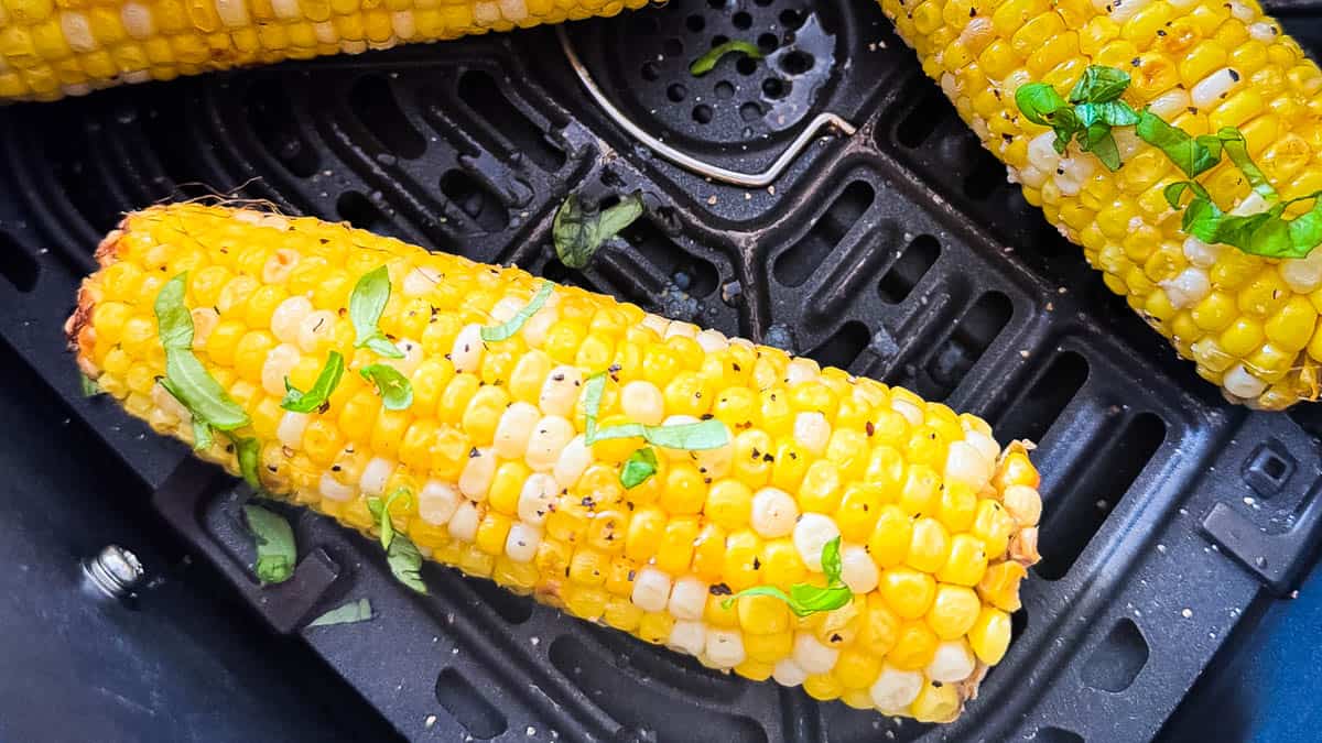 Cooked corn on the cob in an air fryer basket with butter, black pepper, and chopped fresh herbs.