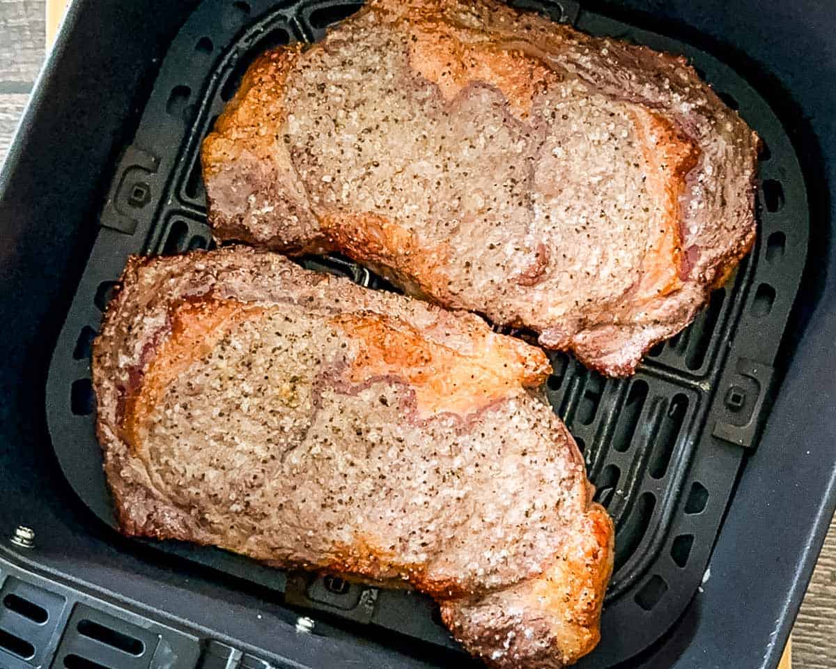 Steaks cooking in an air fryer basket halfway through the cooking process before flipping.