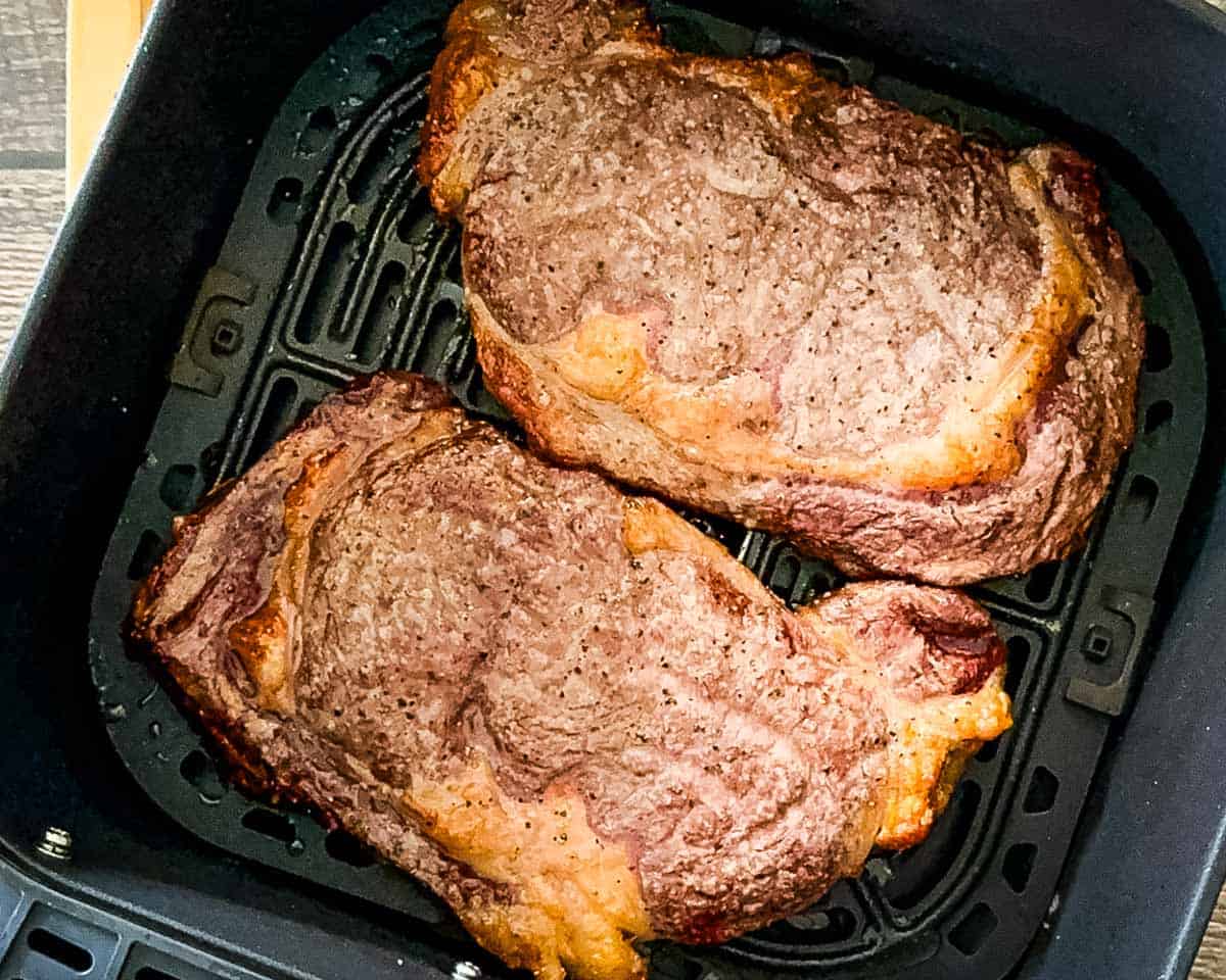 Air fryer ribeye steaks after flipping, showing light browning and rendered fat.