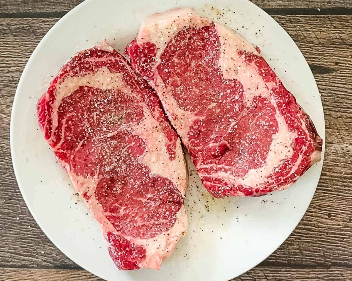 Raw ribeye steaks seasoned with salt, pepper, and spices on a white plate before air frying.
