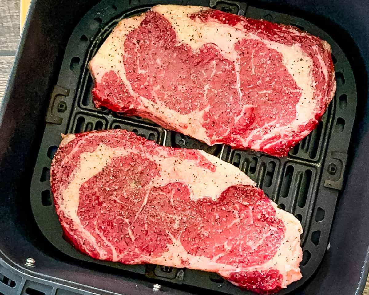 Seasoned ribeye steaks arranged in a single layer in an air fryer basket before cooking.