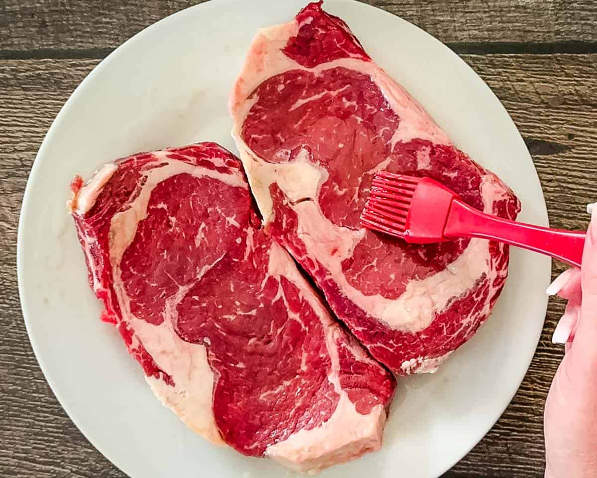Brushing avocado oil onto thick-cut ribeye steaks before seasoning for air fryer cooking.