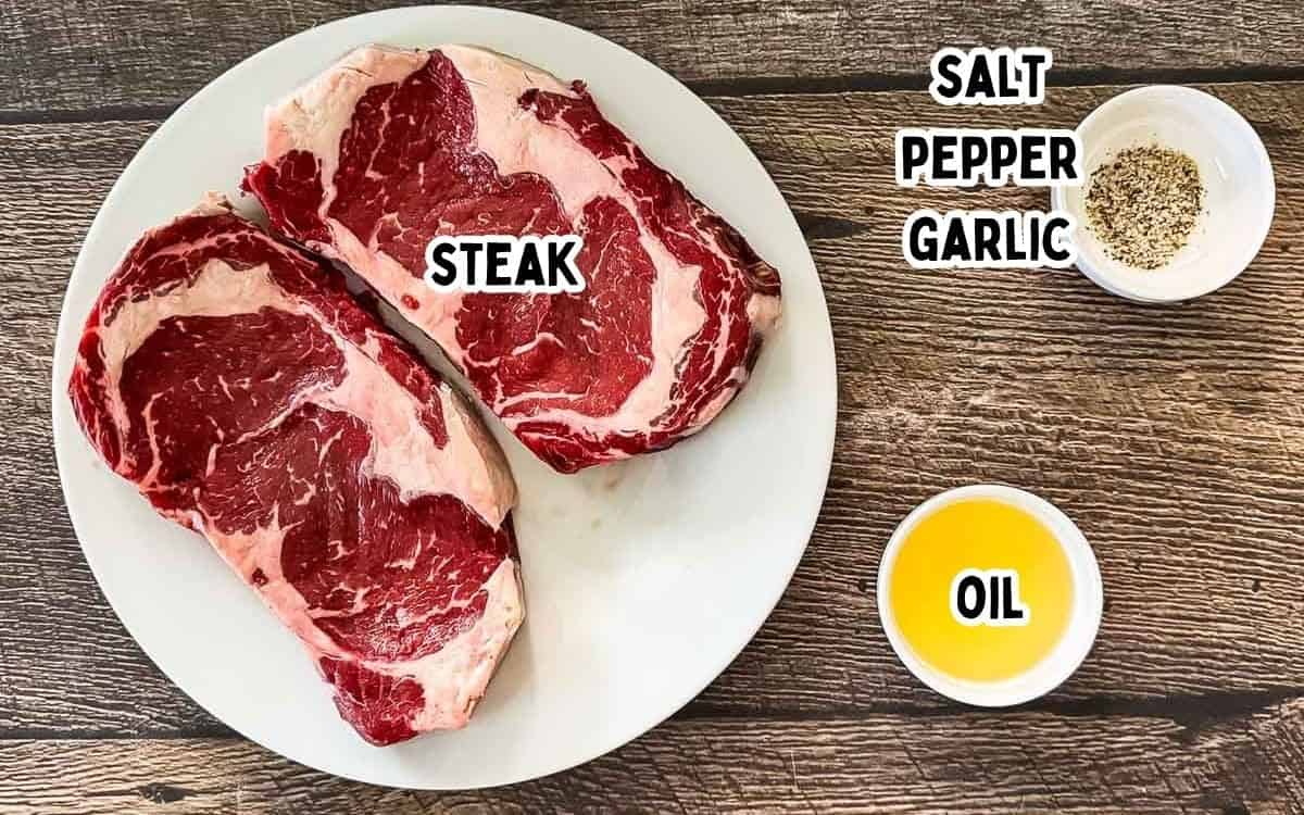 Labeled ingredients for air fryer steak on a wooden table, including steak, salt, pepper, garlic, and oil.