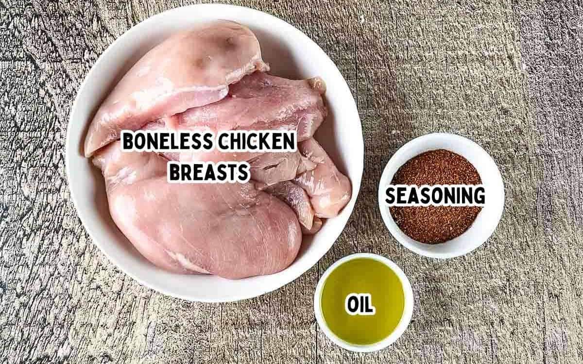 Labeled ingredients for smoked chicken breast in electric smoker, including raw chicken breasts with olive oil and Cajun seasoning on a wood surface.