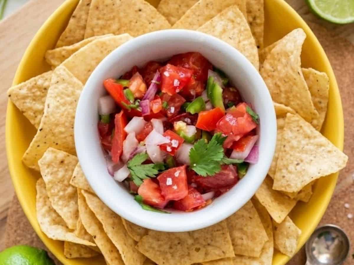 A crisp white bowl filled with fresh keto pico de gallo, nested perfectly in the center of a large yellow bowl overflowing with crunchy tortilla chips.
