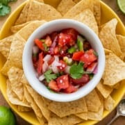A crisp white bowl filled with homemade keto pico de gallo, nested perfectly in the center of a large yellow bowl overflowing with crunchy tortilla chips.