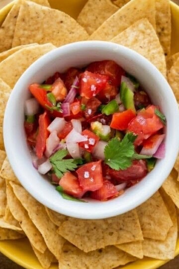 A crisp white bowl filled with homemade keto pico de gallo, nested perfectly in the center of a large yellow bowl overflowing with crunchy tortilla chips.