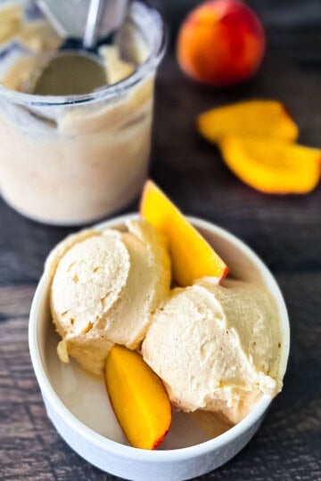 Two scoops of creamy Ninja Creami peach ice cream in a white bowl, topped with fresh peach slices.