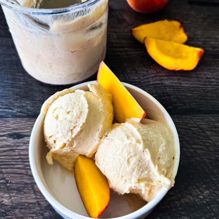 Two scoops of creamy Ninja Creami peach ice cream in a white bowl, topped with fresh peaches.