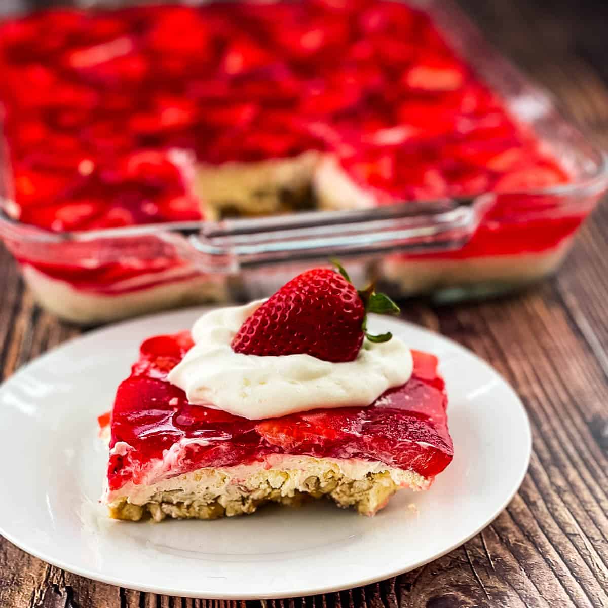 Slice of strawberry pretzel salad topped with whipped cream and a fresh strawberry on a white plate.