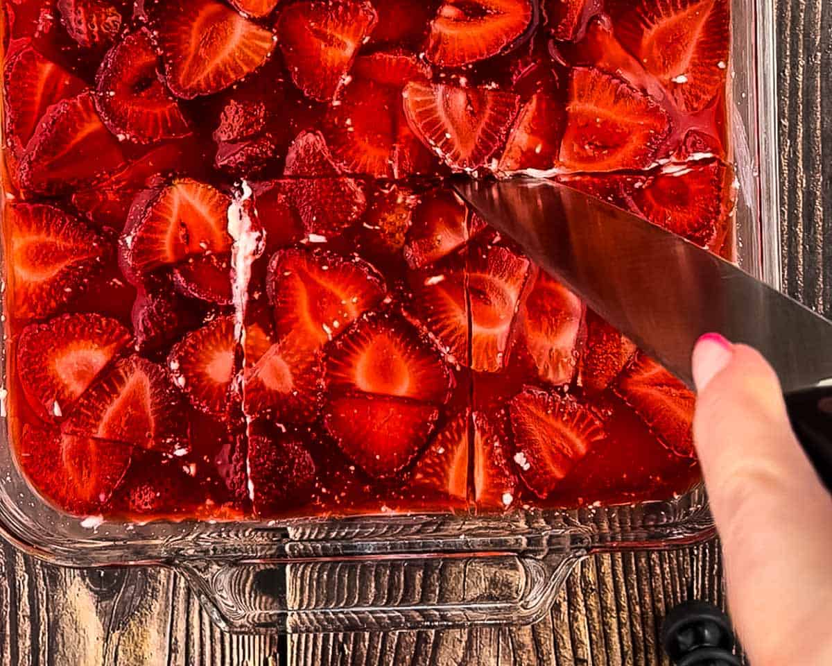 Using a knife to cut a square of strawberry pretzel salad from a glass baking dish with a spatula.