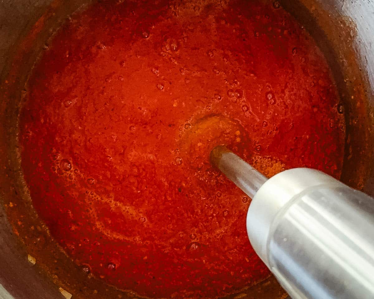 Blending homemade tomato sauce with an immersion blender until smooth.