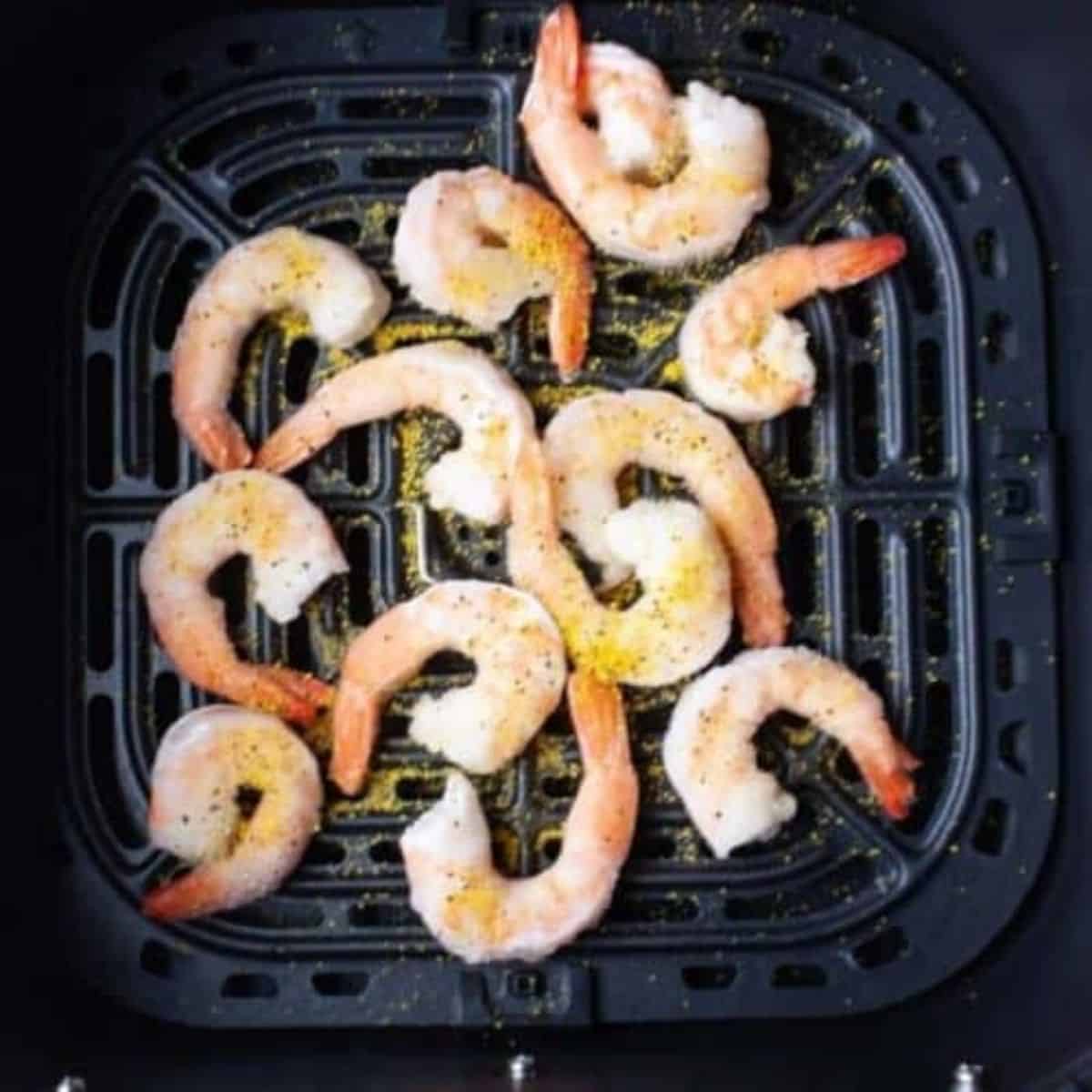 Frozen shrimp in air fryer basket with lemon pepper seasoning.