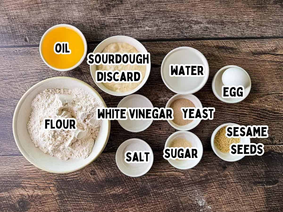 Ingredients needed to make sourdough discard hamburger buns, including flour, active dry yeast, sourdough discard, water, oil, sugar, salt, egg, white vinegar, and sesame seeds.