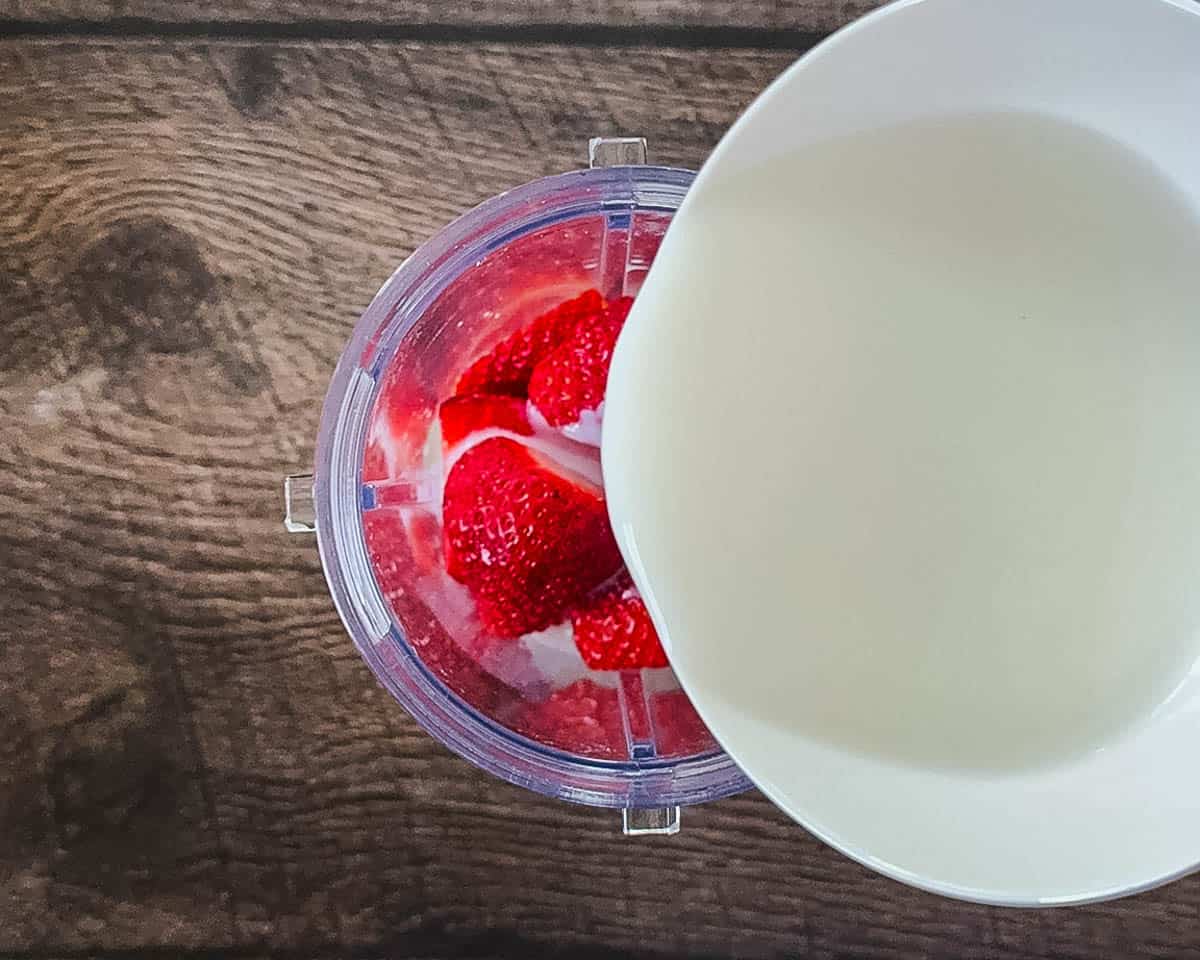 Overhead view of whole milk and heavy cream being poured from a white bowl into a blender jar filled with fresh, bright red strawberries.
