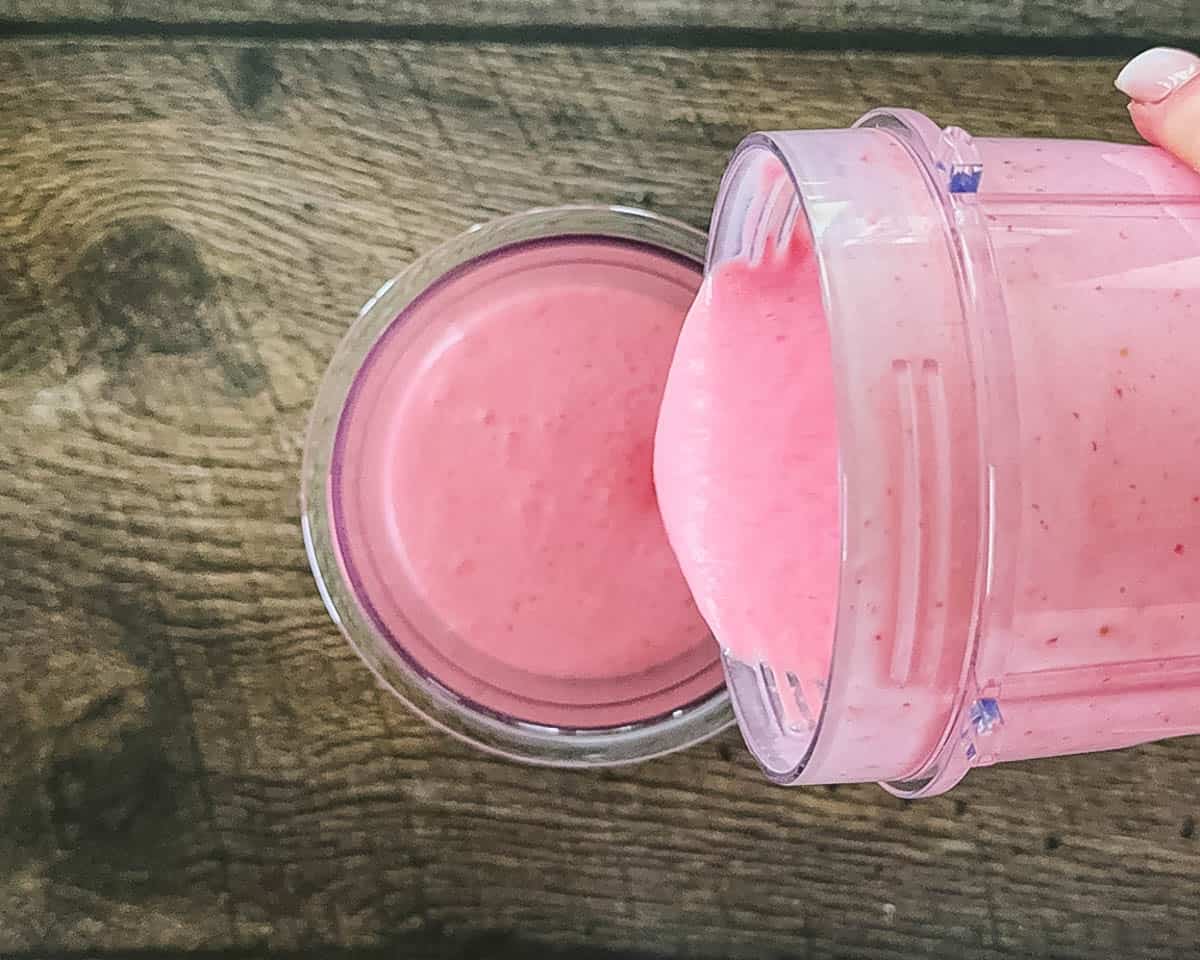 Pouring a smooth, completely blended pink strawberry ice cream liquid base from a blender cup into a clear Ninja Creami pint container.