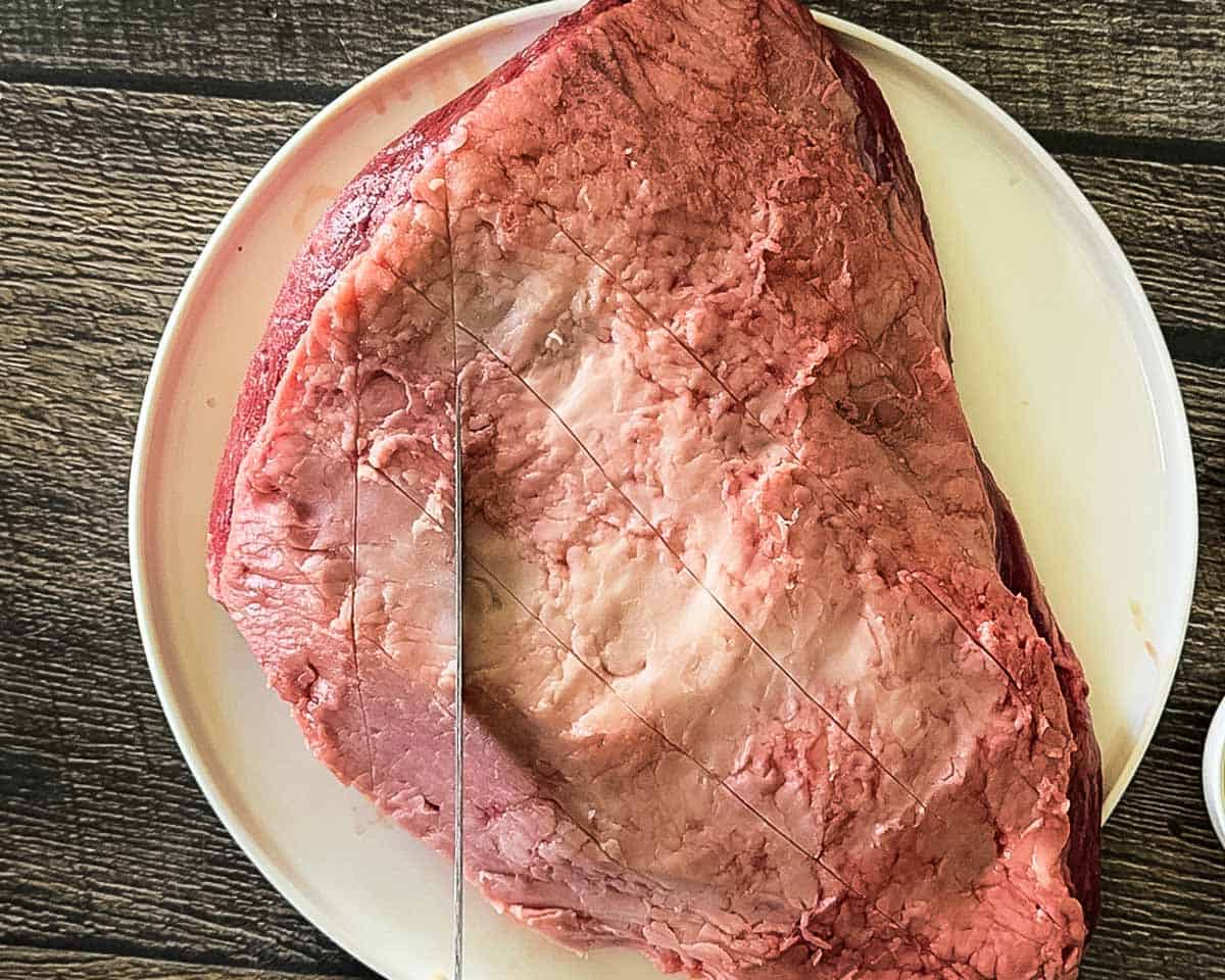 A knife scoring a diamond pattern in a raw trimmed sirloin cap roast (picanha) on a plate before seasoning.