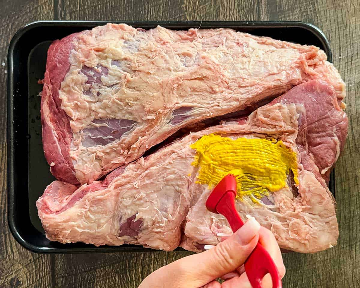 Brushing yellow mustard over two trimmed tri-tip roasts before adding the dry rub.