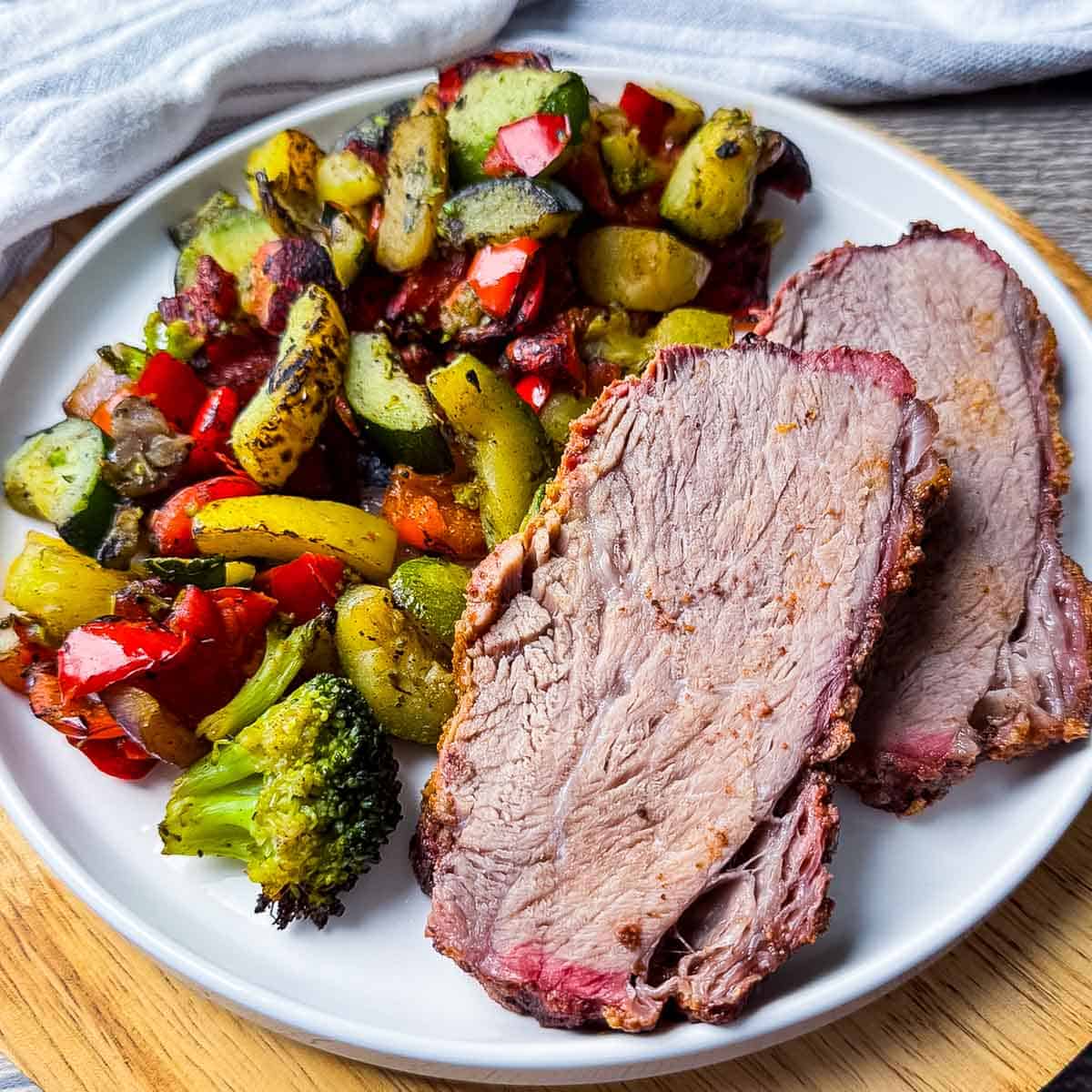 Sliced smoked trisket served with grilled mixed vegetables on a white dinner plate.