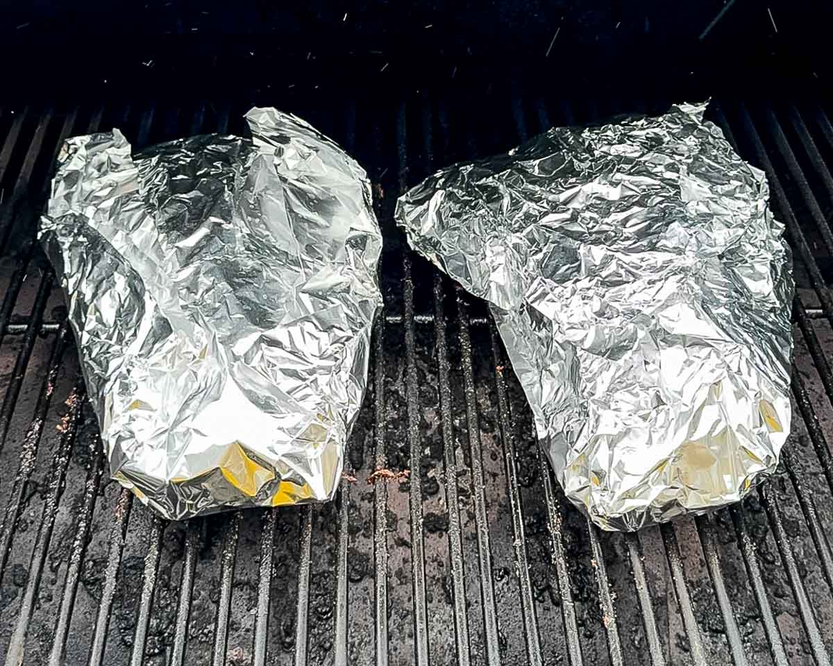 Two foil-wrapped tri-tip roasts cooking on a pellet grill after being wrapped with beef broth and butter.