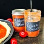 Open and sealed jars of homemade canned pizza sauce with fresh tomatoes beside homemade pizza dough.