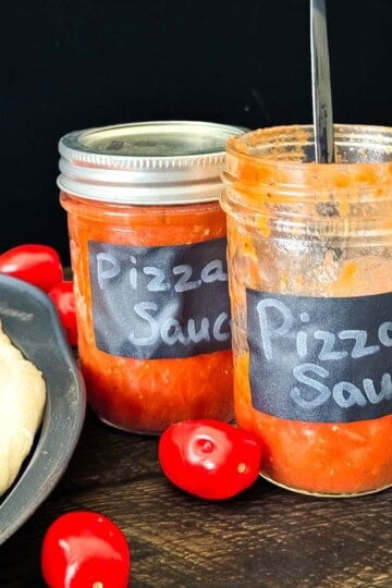 Open and sealed jars of homemade canned pizza sauce with fresh tomatoes beside homemade pizza dough.
