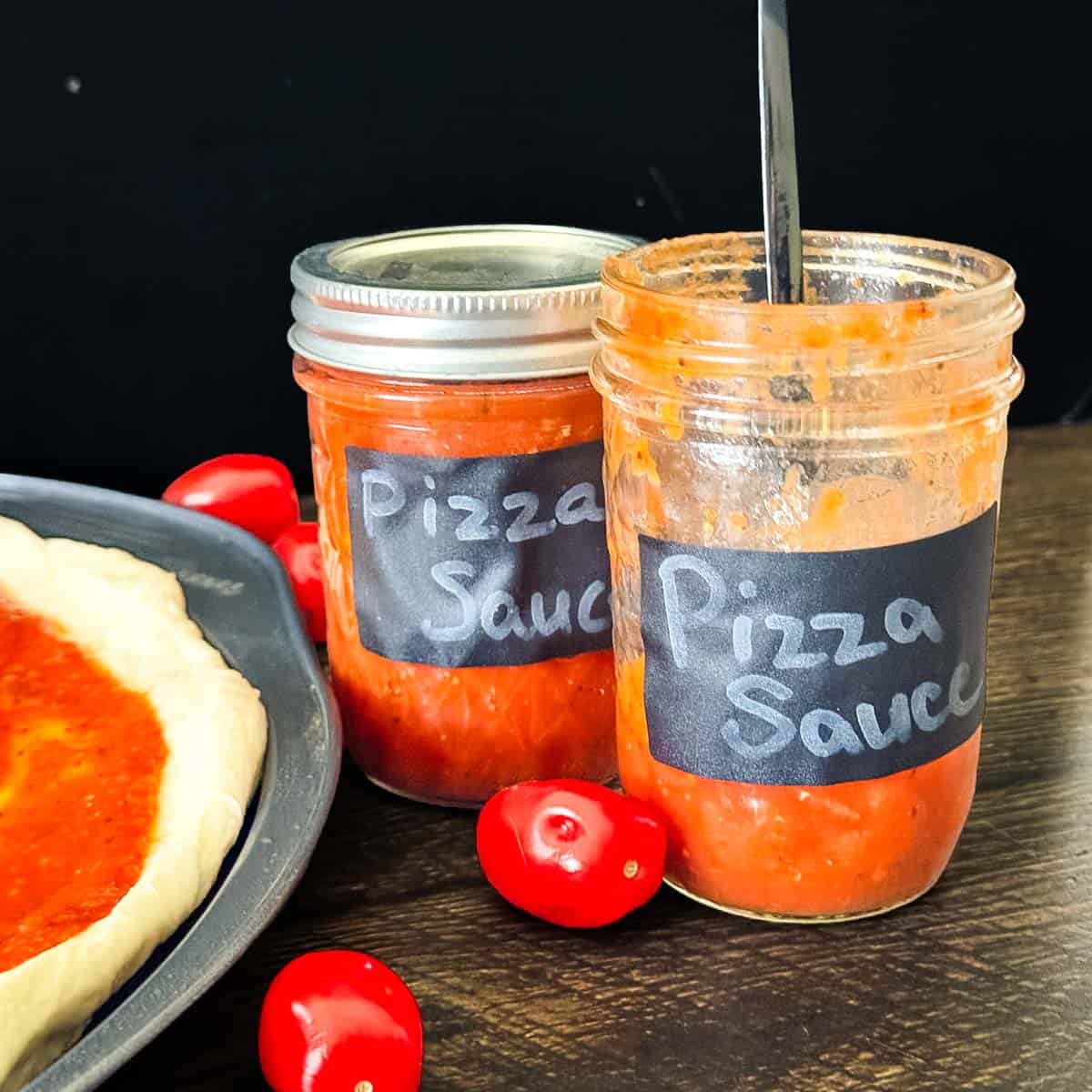 Open and sealed jars of homemade canned pizza sauce with fresh tomatoes beside homemade pizza dough.