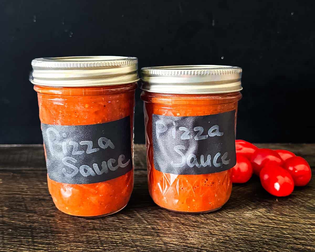 Two sealed jars of homemade canned pizza sauce on a wooden table with fresh tomatoes.