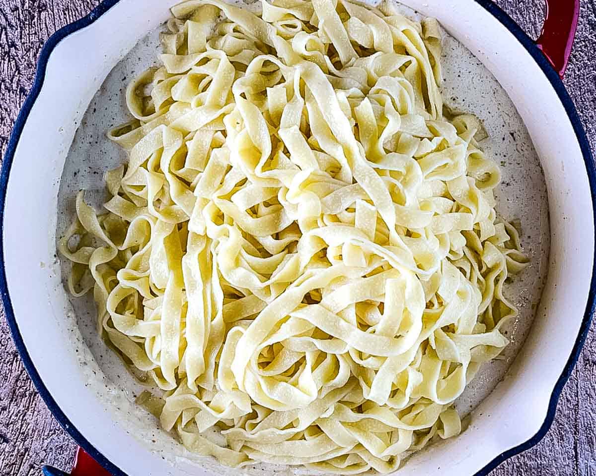 Cooked fettuccine added to creamy Parmesan Alfredo sauce in a skillet.