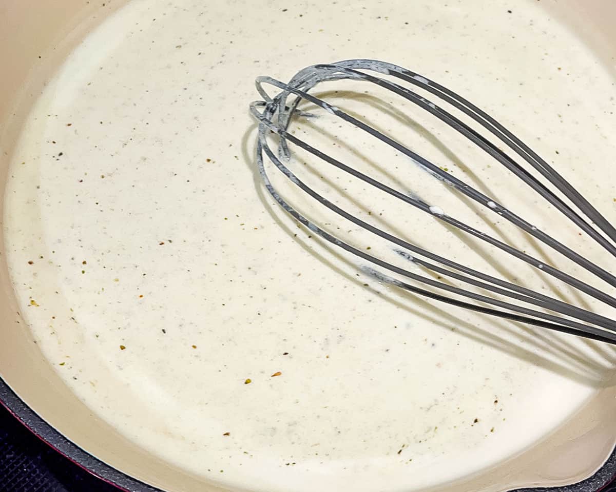 Creamy Alfredo sauce seasoned with Italian seasoning, salt, and black pepper in a skillet.