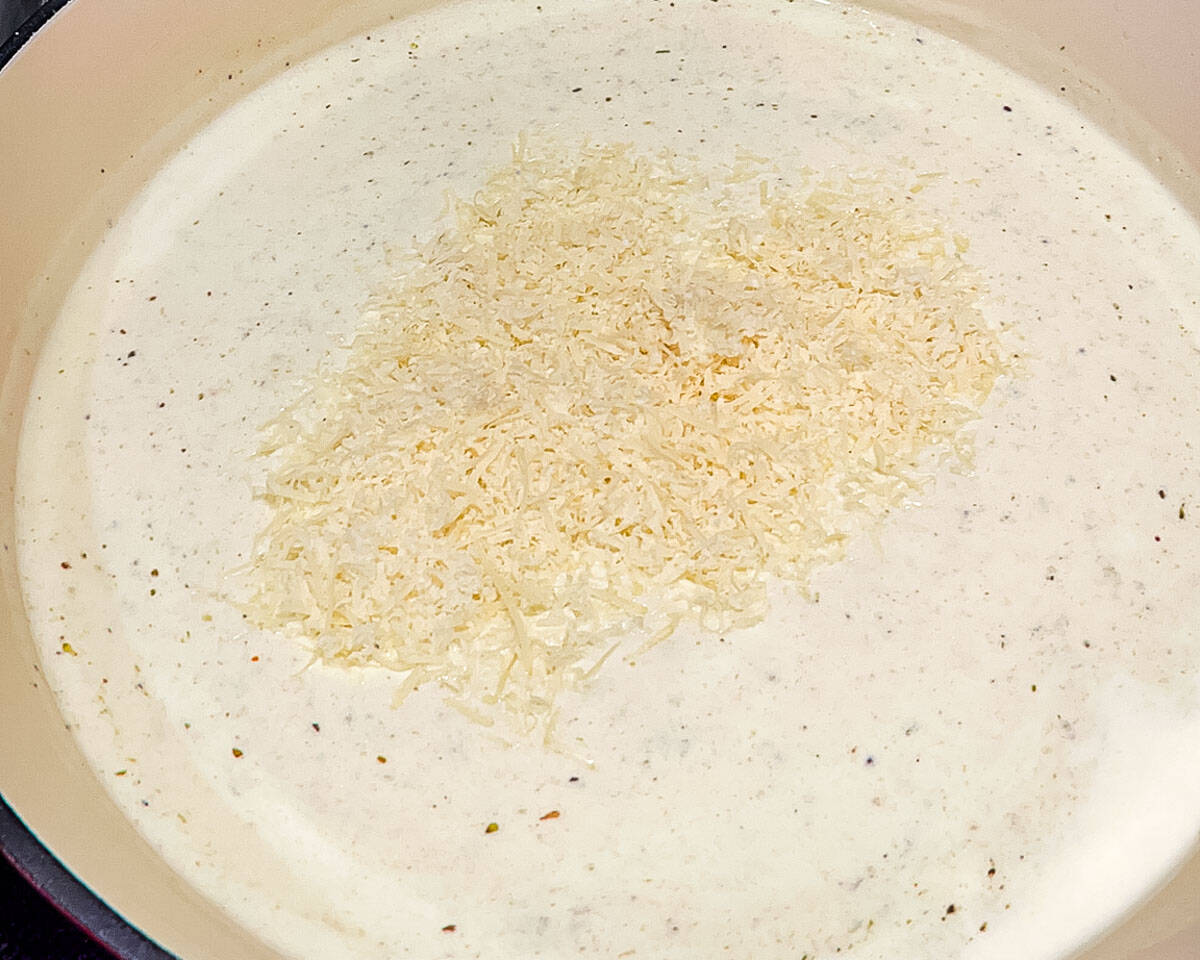 Freshly grated Parmesan cheese added to creamy Alfredo sauce in a skillet.