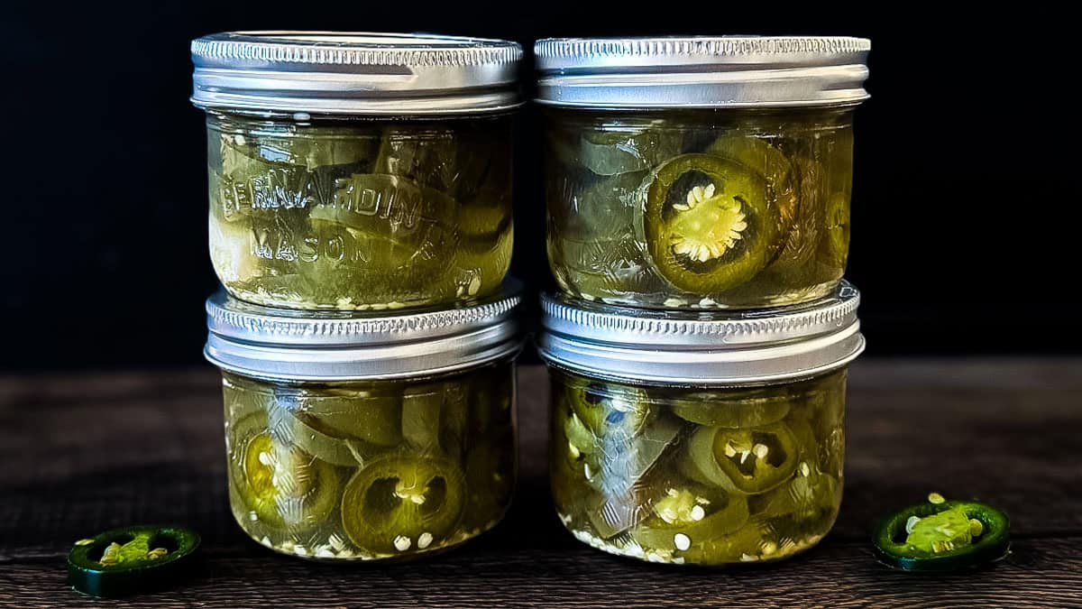 Four small mason jars filled with homemade canned jalapeño pepper slices in pickling brine, stacked on a dark wooden surface with fresh jalapeño slices.