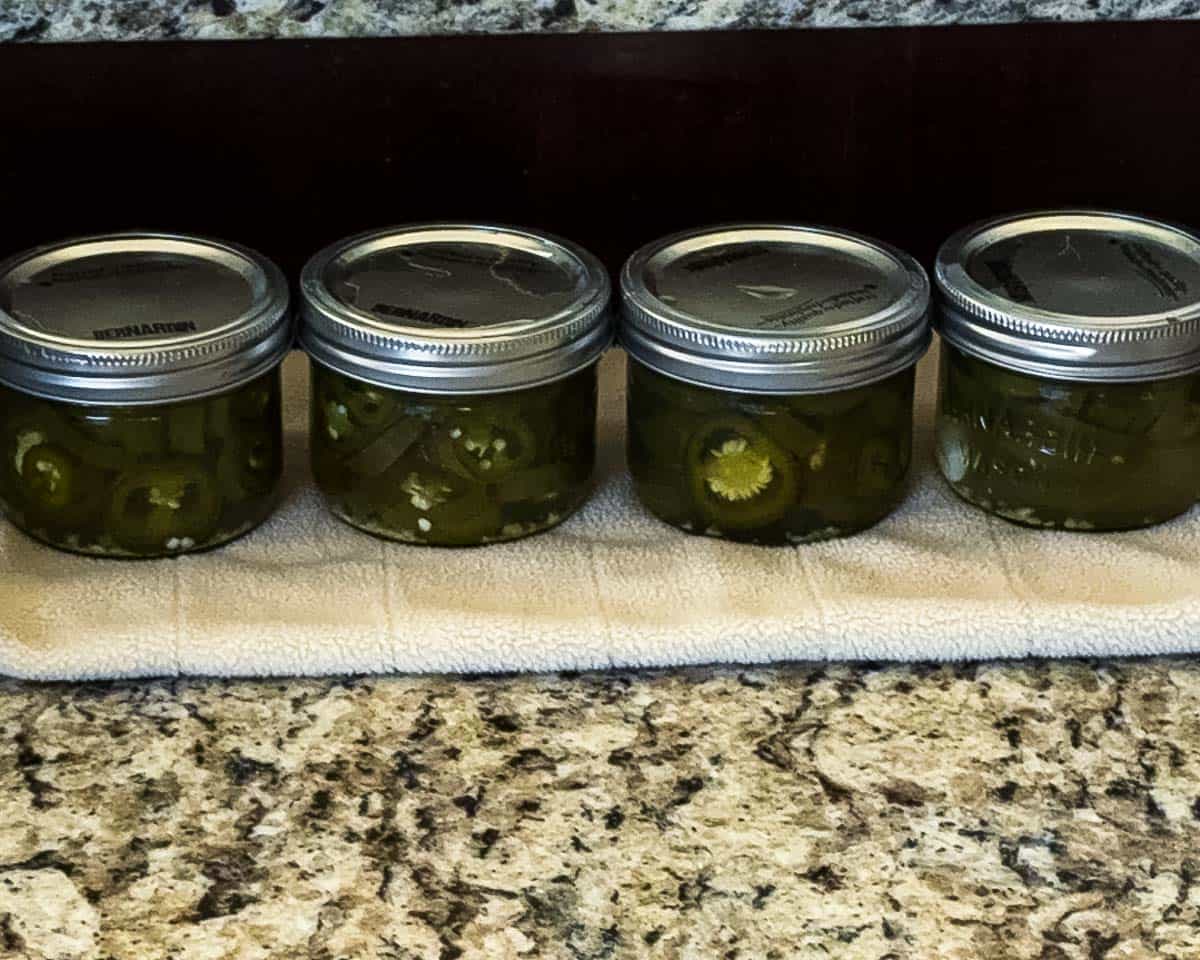 Four jars of homemade canned jalapeño peppers cooling on a towel after water bath canning.