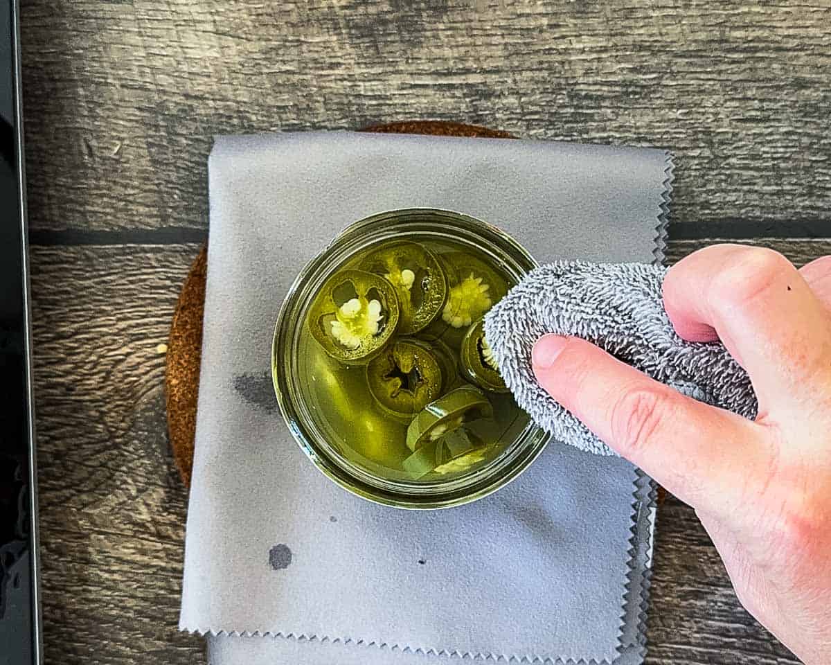 Wiping the rim of a mason jar filled with pickled jalapeño peppers before adding the canning lid.
