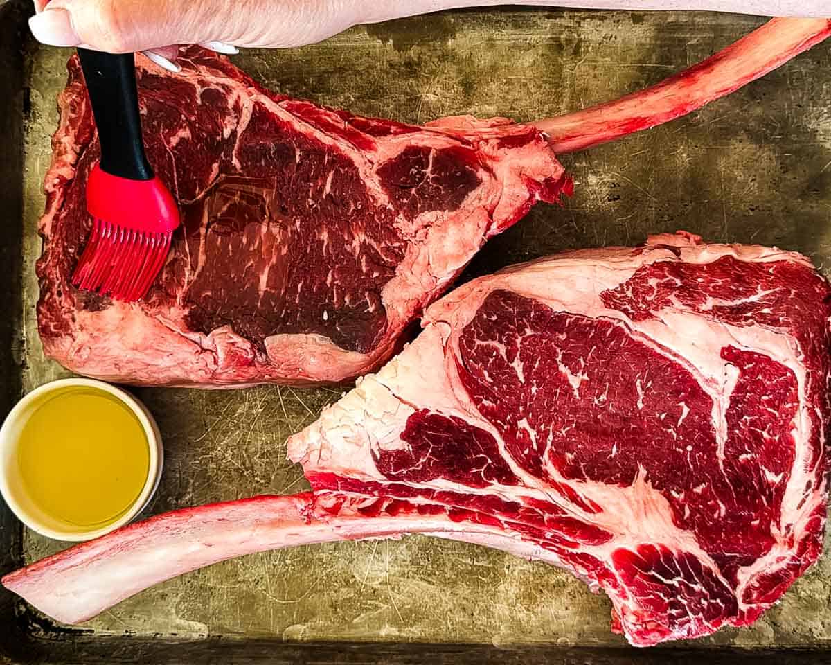 Brushing avocado oil over raw tomahawk steaks before seasoning.