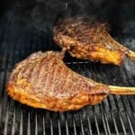 Two smoked tomahawk steaks with long bone handles resting on a smoker grill grate, seared crust developing with visible grill marks and smoke rising.