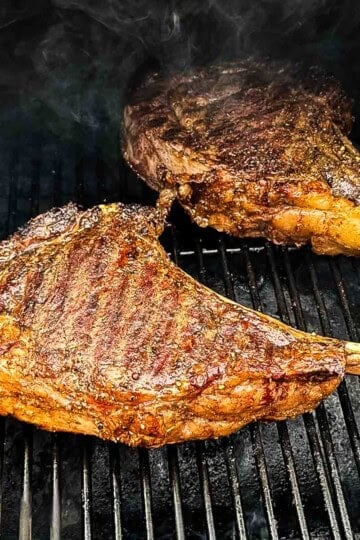 Two smoked tomahawk steaks with long bone handles resting on a smoker grill grate, seared crust developing with visible grill marks and smoke rising.