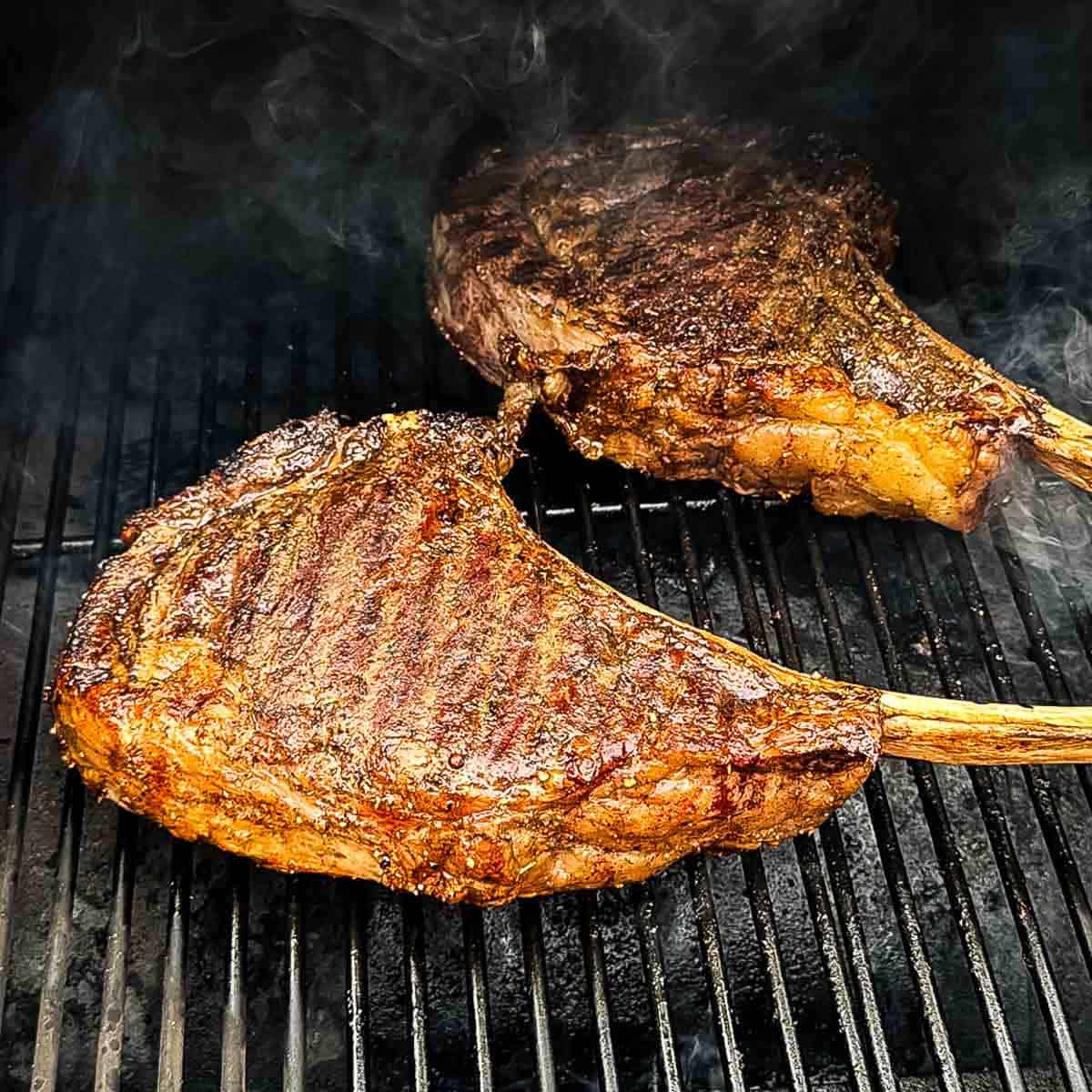 Two smoked tomahawk steaks with long bone handles resting on a smoker grill grate, seared crust developing with visible grill marks and smoke rising.