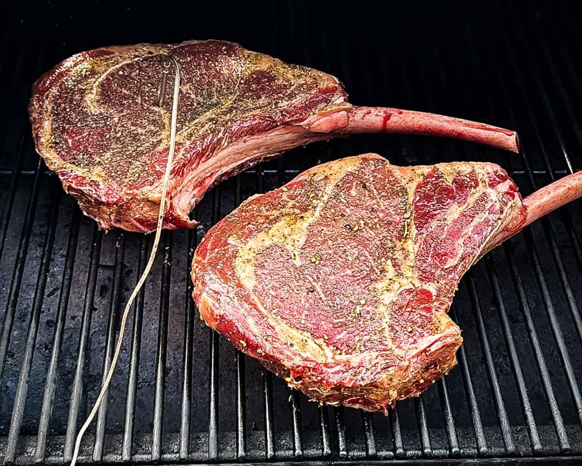 Seasoned tomahawk steaks smoking on a pellet grill with a temperature probe inserted.