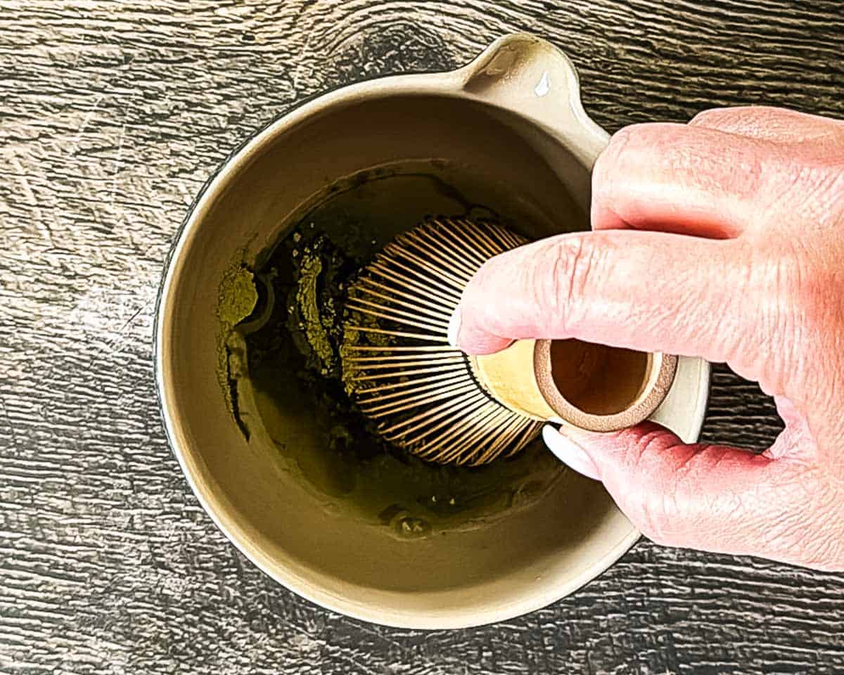Matcha powder and hot water being whisked with a bamboo matcha whisk.