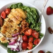 Grilled chicken salad in a large white bowl with strawberries, spinach, and red onion.