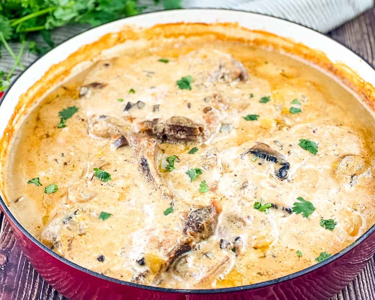 Baked cream of mushroom pork chops in a red cast iron pan with golden, bubbling mushroom gravy and fresh herbs.
