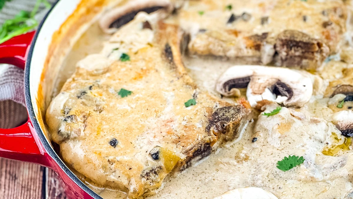 Baked bone-in pork chop in creamy mushroom gravy with sliced mushrooms for garnish in a red cast iron skillet.