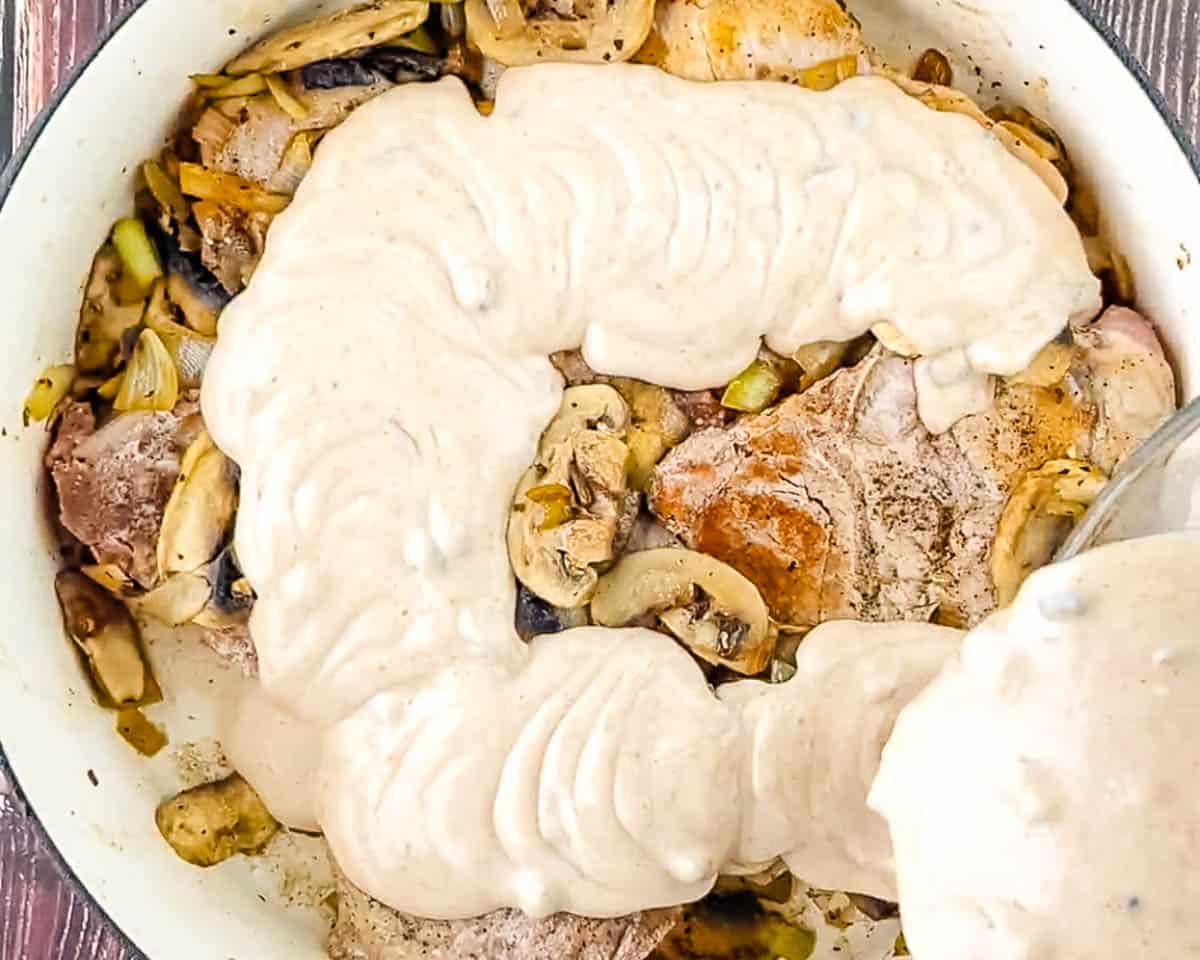 Creamy mushroom soup sauce being poured from a glass bowl over seared pork chops, mushrooms, and onion in a cast iron skillet.