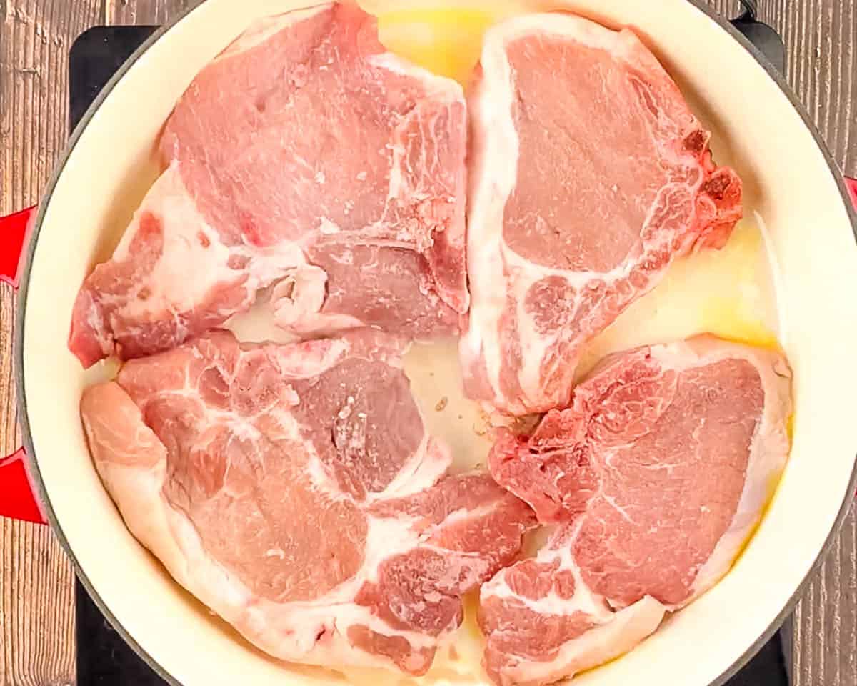 Four raw bone-in pork chops in a cast iron skillet with olive oil, ready to sear.