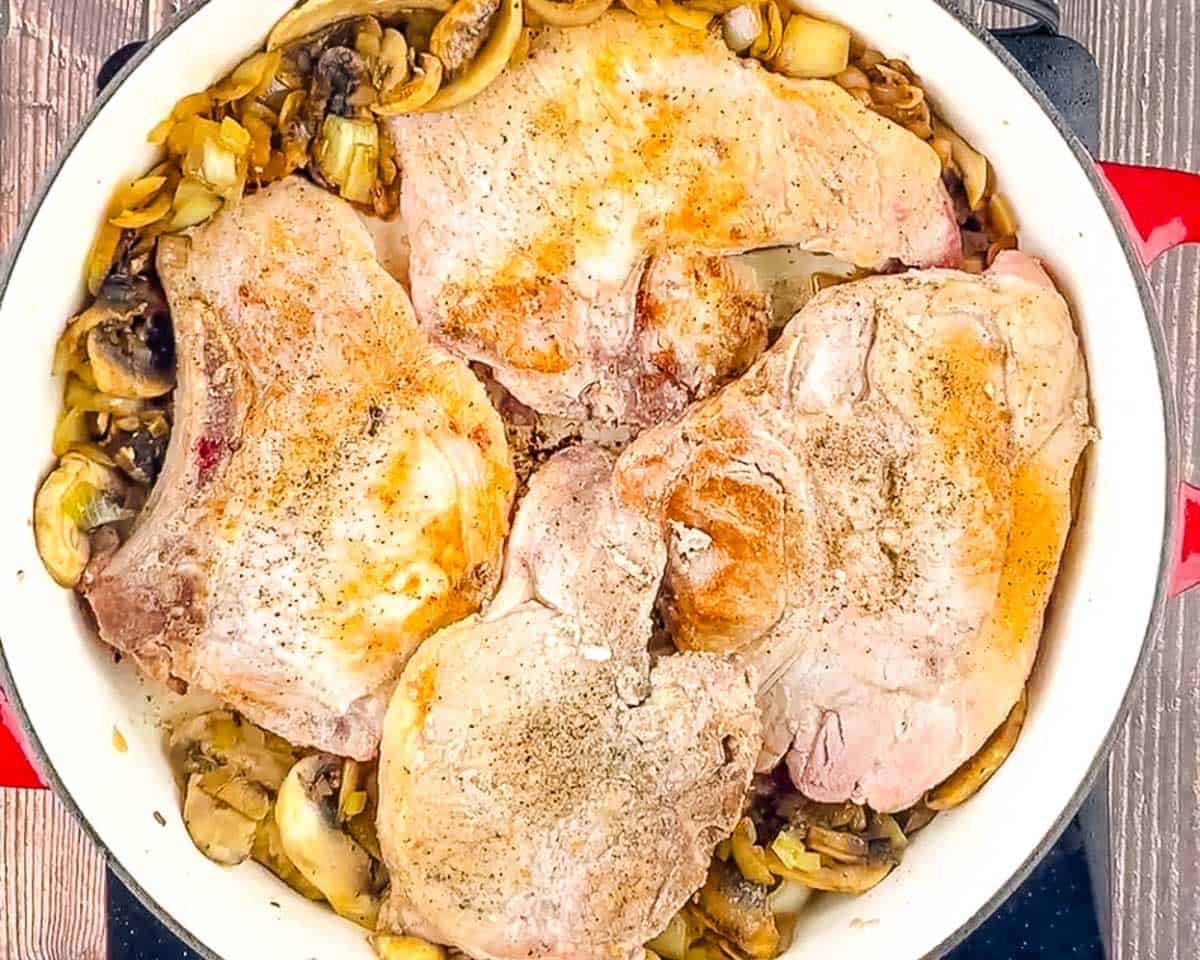 Four seared bone-in pork chops nestled on top of sautéed mushrooms and onion in a cast iron skillet.
