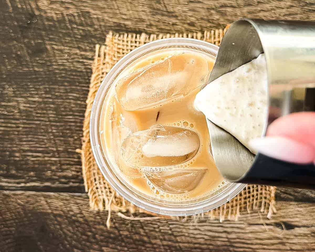 Pouring pumpkin cold foam from a steel pitcher over an iced chai latte.